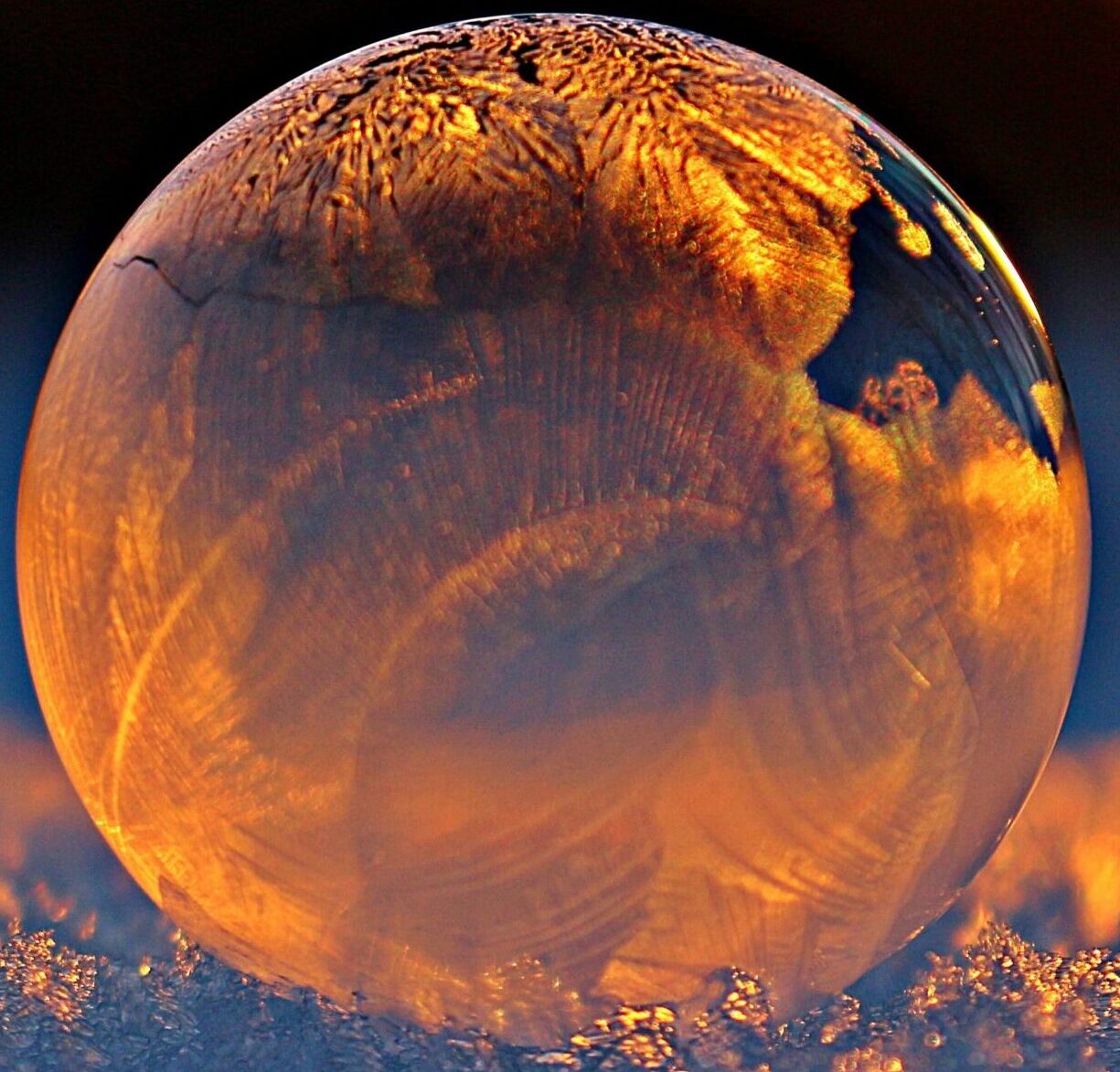 Close-up shot of a frozen bubble with warm reflections resting on a snowy surface at twilight.